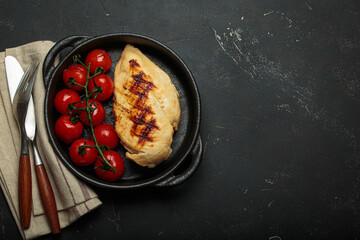 Grilled chicken breast in a cast iron pan on rustic black background, surrounded by roasted cherry tomatoes and cutlery, healthy meal concept, copy space