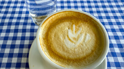 Creamy latte art in white cup on blue plaid table invites hygge mornings, reminiscent of International Coffee Day and Danish coziness