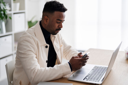 Side view of serious Arab male remote worker messaging on cellphone while sitting at table with netbook in modern apartment during workday
