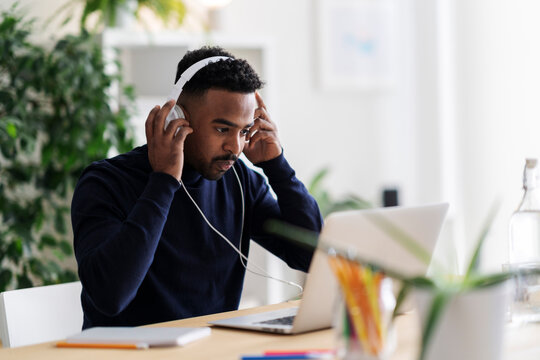 Young Arab male self employed remote worker sitting at table with laptop and notepad and putting on headphones while working in modern workspace at home