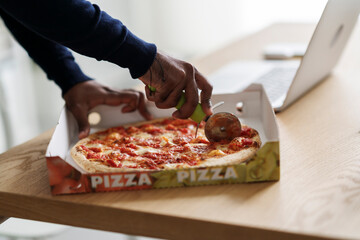 High angle of crop anonymous Arab male slicing tasty delivered pizza in box placed on table near laptop for distant work from convenient home office