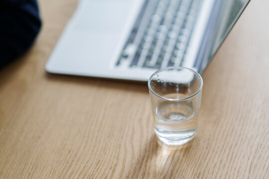 High angle of transparent crystal glass of fresh water placed near modern netbook on desk at workplace for remote project