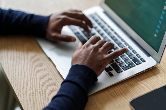 High angle of crop anonymous Arab male freelancer sitting at wooden table and typing on netbook while doing online job at convenient workplace