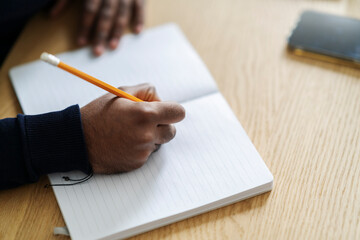 High angle of crop anonymous Arab male writing ideas in planner at wooden table with smartphone at daylight