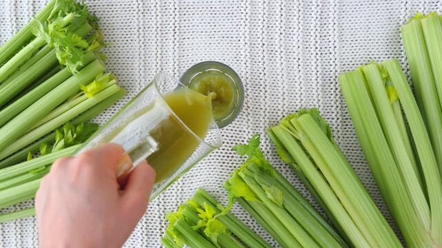 Top view of a hand pouring freshly squeezed celery juice from a pitcher into a glass, surrounded by green celery stalks on a white knitted background, illustrating a healthy diet and detoxification