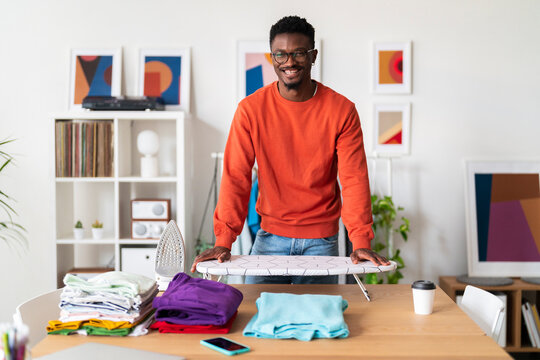 Smiling black man in casual outfit standing near table with folded clothes after ironing in modern living room