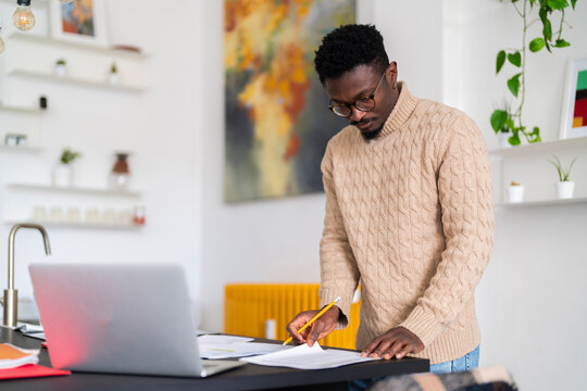 Concentrated young black bearded man in eyeglasses and casual clothes standing near table with laptop while checking bills