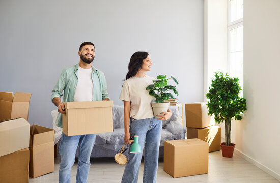 Happy couple moving with boxes into new home apartment. Young man and woman smile while unpacking in living room, homeowners holding plant and lamp, standing together by cardboard boxes pile and sofa - Powered by Adobe