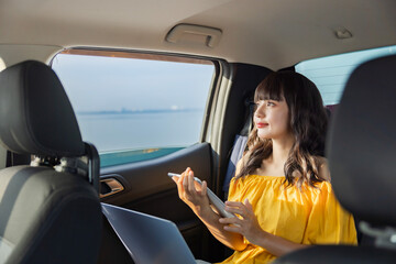 Naklejka premium Car insurance, Technology concept - Confident Woman working on laptop and using tablet search hotel, online shopping at back sit of car on a bright day with blue sky sunlight. Happy along the way trip