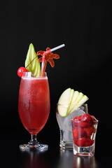 A refreshing red cocktail with garnish, ice, and cherries, against a dark backdrop. The glass is garnished with slices of green apple and cherry, offering a visually appealing and refreshing drink.