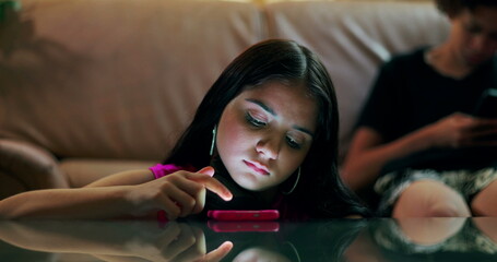 Teenager girl engrossed in smartphone on table with glowing light on her face, boy in background...