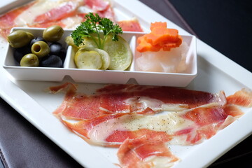 A close-up shot of a gourmet platter with various delicious food items. The platter includes olives, pickles, cured meats, and other appetizers