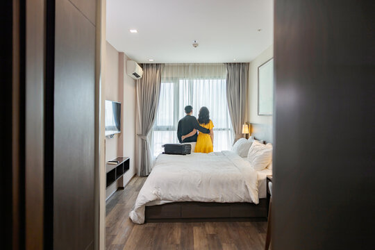 Back side photo of nice asian couple hugging each other standing nearly window, looking to beautiful view outside in hotel/resort bedroom after check-in or moving in to new house or apartment.