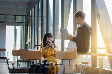 Happy traveler using digital tablet while his girlfriend is using cellphone at airport terminal. Travel, airport and terminal for departure or arrival with boarding gate.