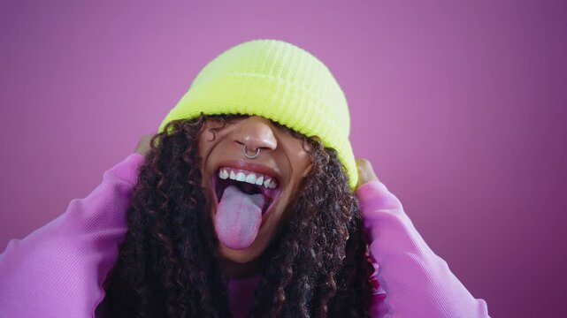 Young woman laughing, pulling fluorescent yellow beanie down over her eyes against a vibrant pink background, playful, confident, colorful street-style headshot conveying joy and authenticity