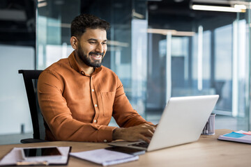 Young indian businessman smiling and typing on a laptop, sitting at a desk in a contemporary office...