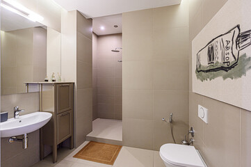 Modern bathroom featuring neutral beige tiles, a white sink and toilet, a walk-in shower, and a minimalist aesthetic. A framed Aesop artwork adds a touch of sophistication