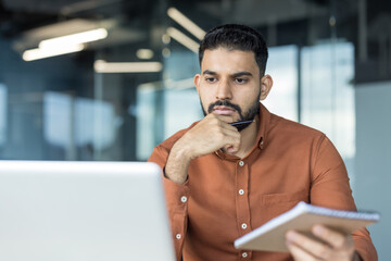 Indian man deeply concentrating while reviewing content on his laptop and holding a pen and notepad, reflecting business strategy or problem-solving in a modern office environment
