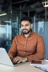 Fotobehang Beren Indian businessman working on a laptop, standing at a desk in a contemporary office setting with glass walls, reflecting a positive attitude and professional success while engaging in business tasks  © Liubomir