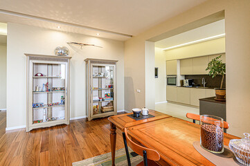 Warm interior features dining area with a wooden table, chairs, and display cabinets. An open doorway leads to a modern kitchen with neutral cabinets. Hardwood floors add to room inviting ambiance