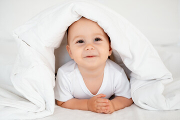 smiling baby girl under a blanket on a white bed, space for text, a small happy child peeking out from under a blanket or going to bed