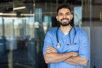 Fototapeta premium Young indian healthcare professional man standing with arms crossed, smiling confidently, wearing blue scrubs and a stethoscope in a modern medical environment