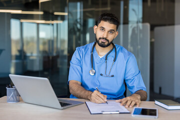 Young male doctor or medical intern wearing blue scrubs and a stethoscope, sitting at a modern office desk, writing on a clipboard with a laptop nearby