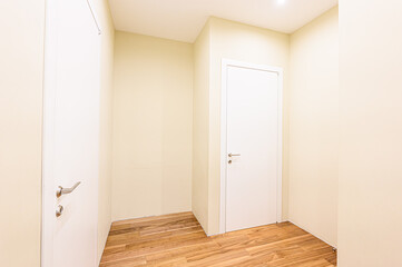 Interior shot of a hallway with two white doors, light yellow walls, and a wooden floor. Bright lighting