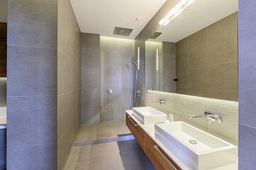 A modern bathroom features sleek grey tiling, a large mirror above a vanity with dual rectangular sinks, chrome faucets, and minimalist lighting. The shower has a large square rain head