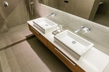 A modern bathroom with a double sink vanity, featuring rectangular white sinks, sleek silver faucets, and wood-tone cabinetry under a large mirror. Neutral color scheme