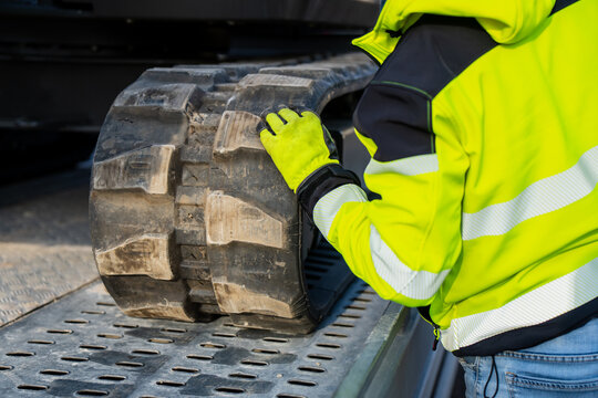 Construction worker in reflective safety jacket handling excavator rubber track on metal platform showing industrial machinery maintenance, transportation process and professional engineering process