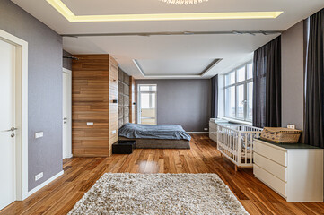 A modern bedroom features a plush bed, a white crib, and wooden accents. Natural light pours in, highlighting the wooden floor