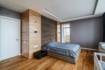 Cozy modern bedroom with a wooden accent wall, a large bed with a gray blanket, and a dark gray wall. A window brings in natural light. Warm wood floor