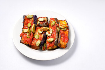 A white plate holds several slices of fried eggplant covered in a spicy red chili sauce, known as balado, and garnished with petai.