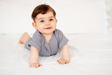 Fototapeta premium smiling baby on a white bed after sleeping, cute baby in a gray cotton bodysuit on a bed, space for text, baby boy