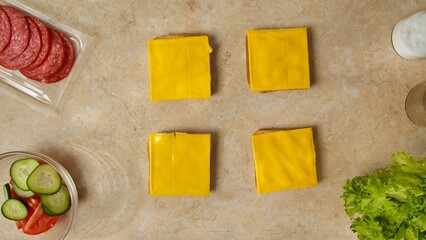 A flat lay of sliced cheese on bread with salami, cucumber, tomato, lettuce, and seasoning on a marble surface. Preparing a healthy breakfast or snack in the kitchen at home.