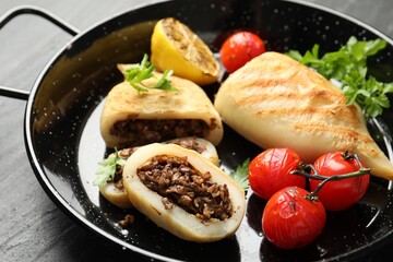 Pieces of tasty grilled squid stuffed with mushrooms, parsley, tomatoes and lemon in pan on dark textured table, closeup