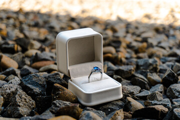 Stunning sapphire engagement ring in a white box placed on rugged dark gravel, symbolizing a bold, outdoor proposal or unique commitment