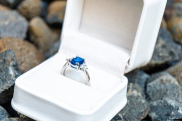 Stunning blue sapphire engagement ring in a white box, set against a backdrop of natural stones. Perfect for luxury and romance themes