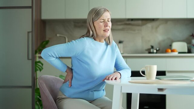 Tired senior gray haired female suffering from back pain sitting at kitchen table at home. Upset elderly retired woman massages painful lower back muscles. She is exhausted or has sciatica