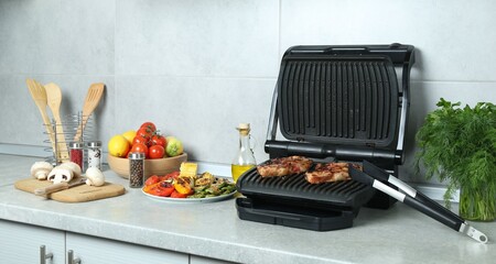 Electric grill with delicious steaks and vegetables on light table in kitchen