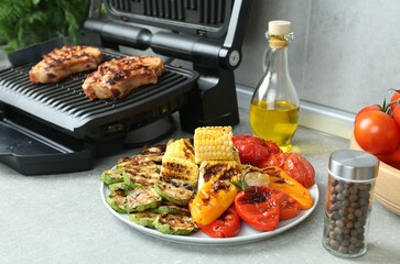 Delicious grilled vegetables and electric grill with steaks on light table, selective focus