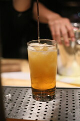 A bartender expertly pours liquid into a tall glass, creating a refreshing drink. The setting is likely a bar, and the drink looks inviting