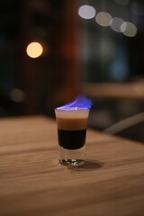 A close-up shot of a meticulously layered B-52 cocktail with a blue flame. The cocktail sits on a wooden table, offering an enticing display of colors and textures.