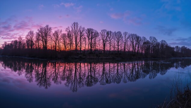 Vibrant evening skyline along a peaceful river with leafless trees mirrored in still waters under a rosy winter sky