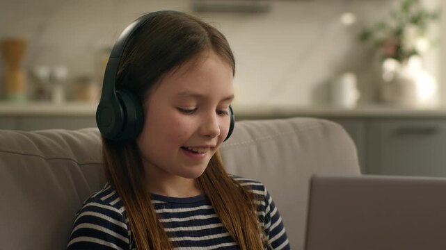 Active happy Caucasian little girl child kid daughter in headphones listening music with laptop computer technology enjoy audio song listen dancing moving sitting on couch dance smiling having fun joy