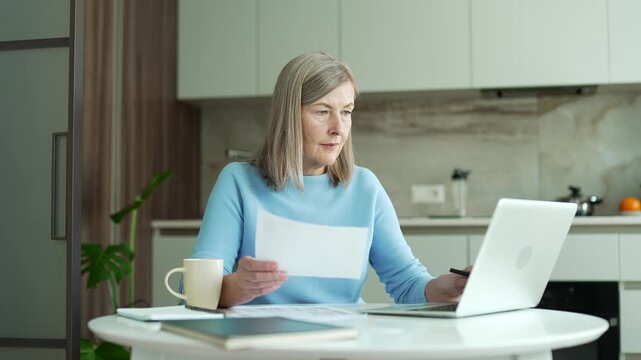 Busy senior gray haired female accountant working doing paperwork sitting at home kitchen. Confident elderly business woman financier makes financial calculations, types on laptop, fills out a form