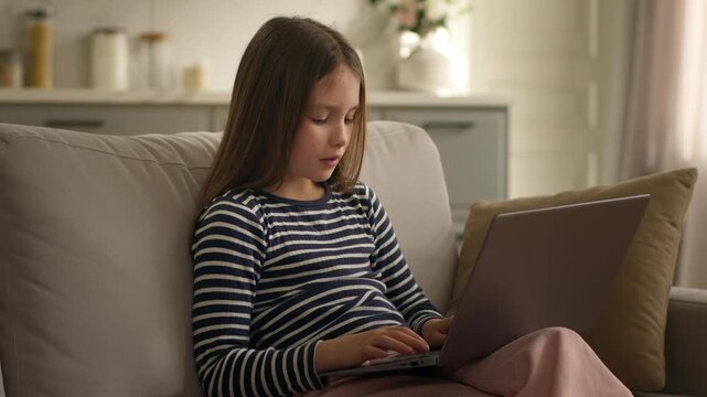 Caucasian little girl child kid daughter schoolgirl pupil using laptop e-learning at home couch typing computer concentrate studying learn with online internet service technology distant education