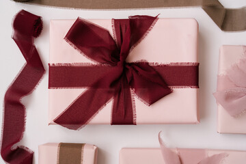 Flat lay of elegant pink gift boxes wrapped with soft fabric ribbons in burgundy and dusty rose shades on a white background. Minimalist aesthetic, cozy atmosphere, pastel tones.