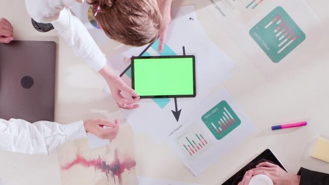 High angle shot of office team reviewing tablet with green screen among charts and coffee cups. Concept of teamwork, digital presentation, cooperation, and focused workflow with analytical energy.